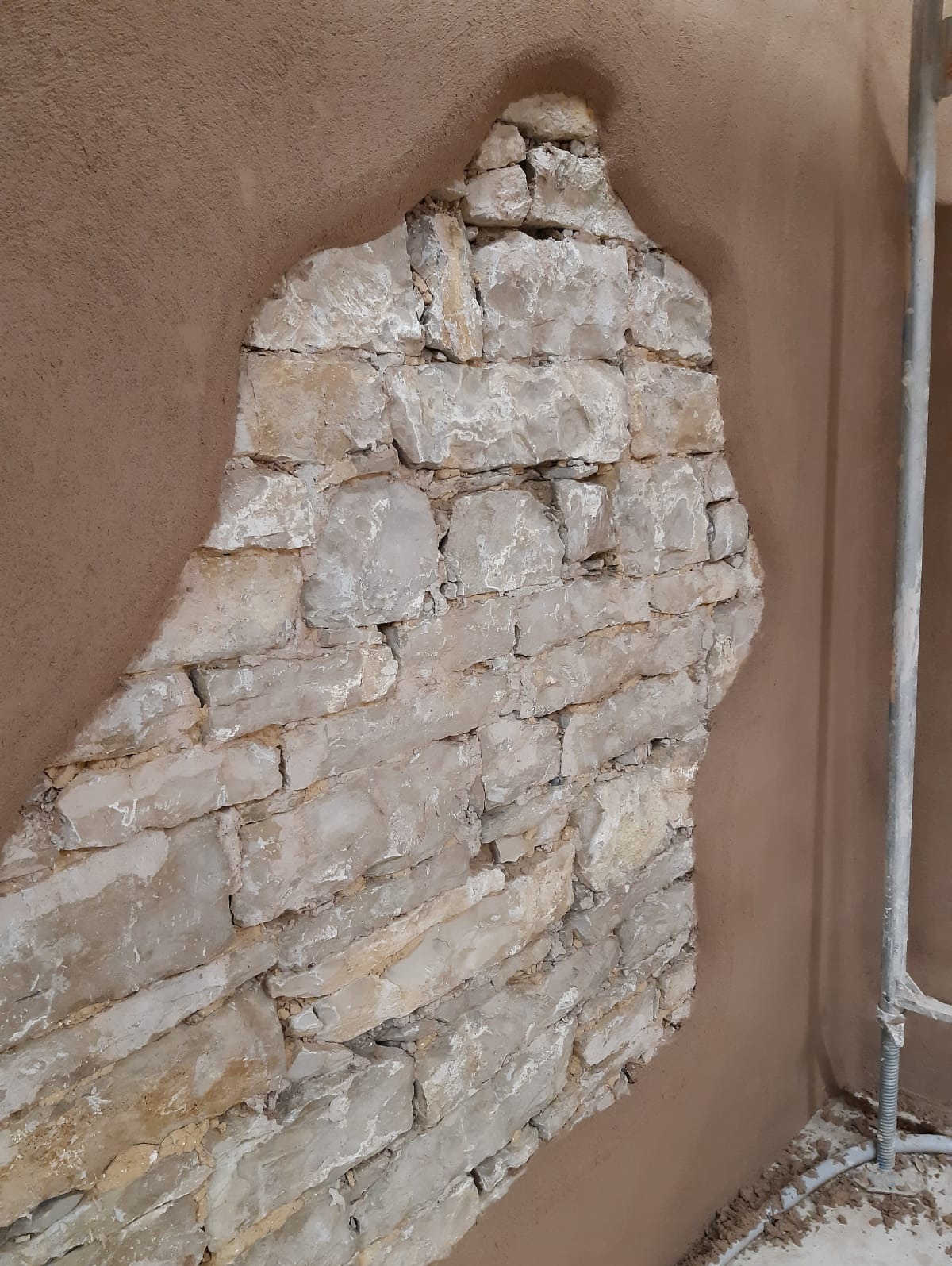 Fragment ściany z kamienia częściowo pokryty świeżym, brązowym tynkiem. Widoczny metalowy stelaż rusztowania.