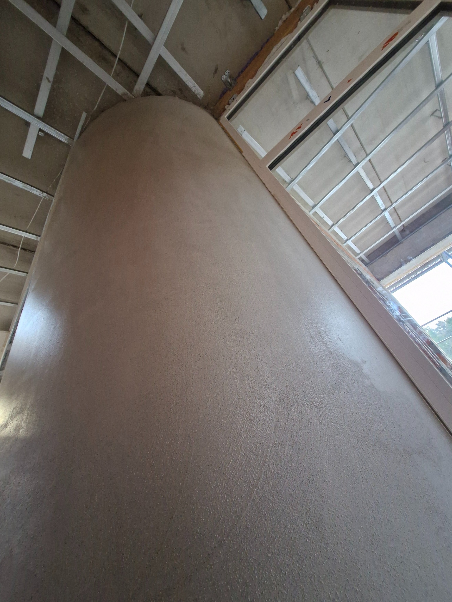 Świeżo nałożona, gładka, beżowa powierzchnia ściany, zakrzywiona u góry, z widoczną konstrukcją sufitu podwieszanego i ramą okienną w tle.