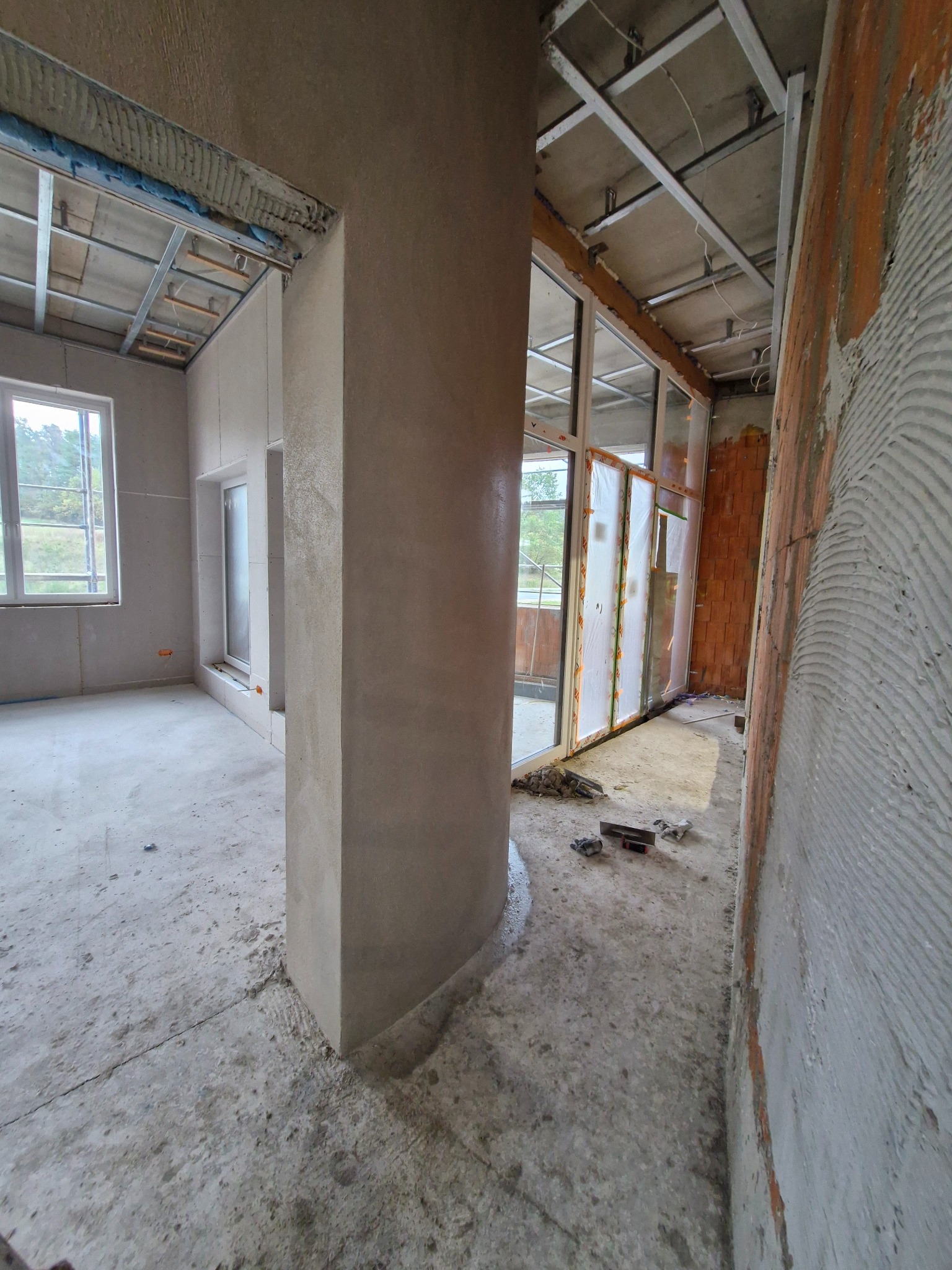 Wnętrze w trakcie prac wykończeniowych; na ścianach surowy tynk, widoczne profile sufitowe, okna zabezpieczone folią, na podłodze resztki materiałów budowlanych i narzędzia.