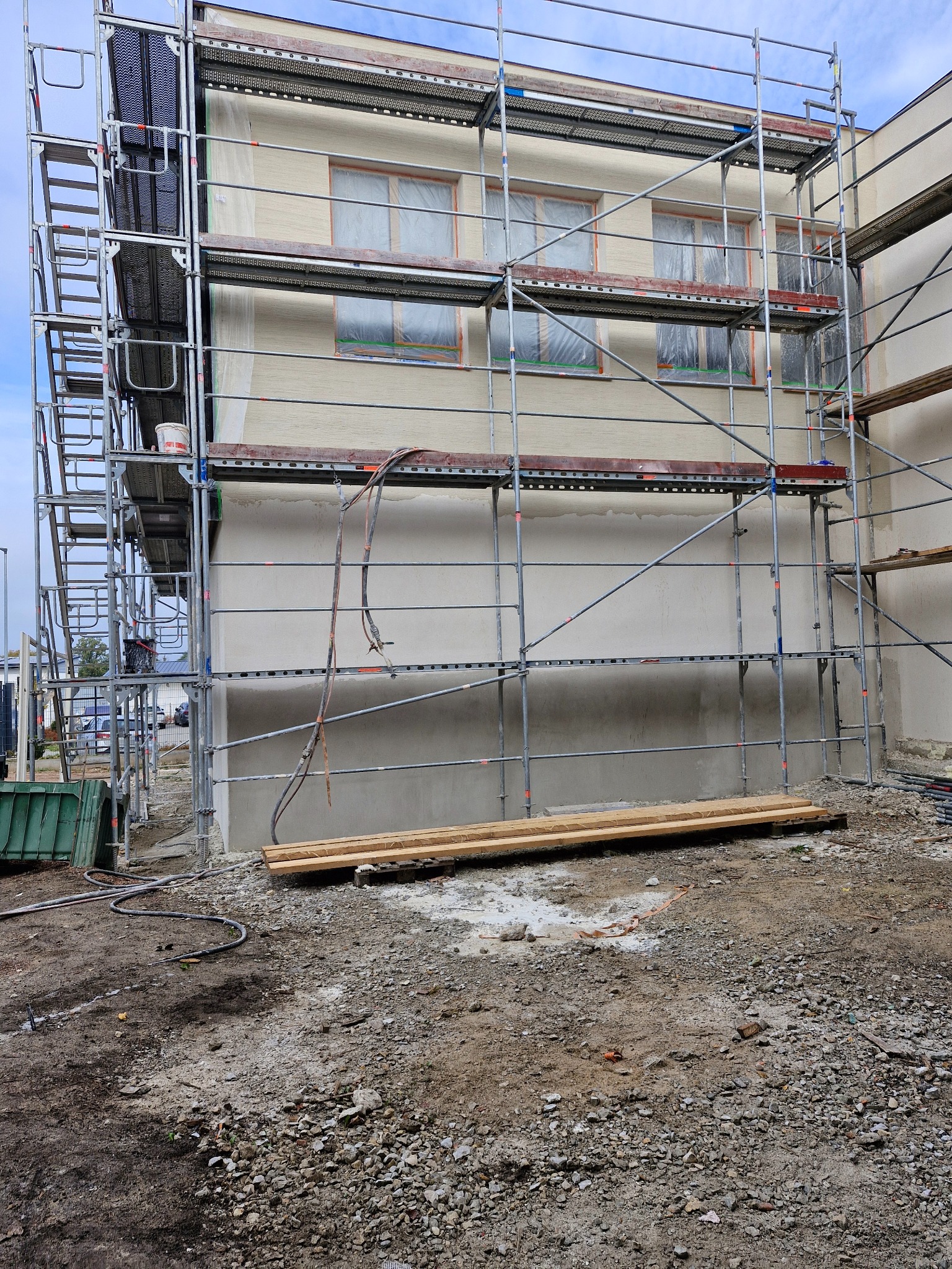 Budynek w trakcie prac elewacyjnych, z rusztowaniem, zabezpieczonymi oknami i widoczną surową warstwą tynku.