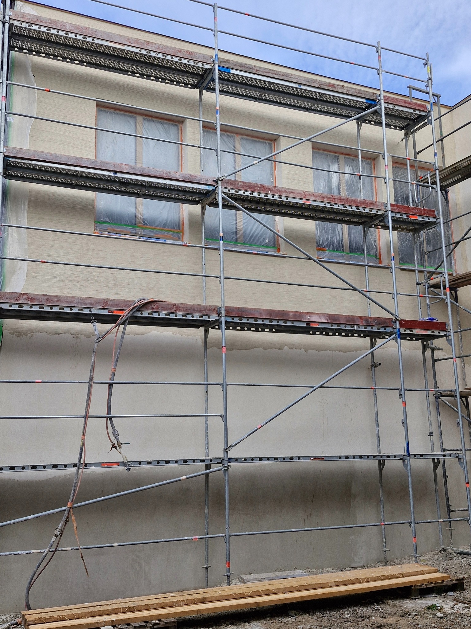 Elewacja budynku w trakcie ocieplania, widoczne rusztowania, okna zabezpieczone folią, częściowo nałożony tynk strukturalny.