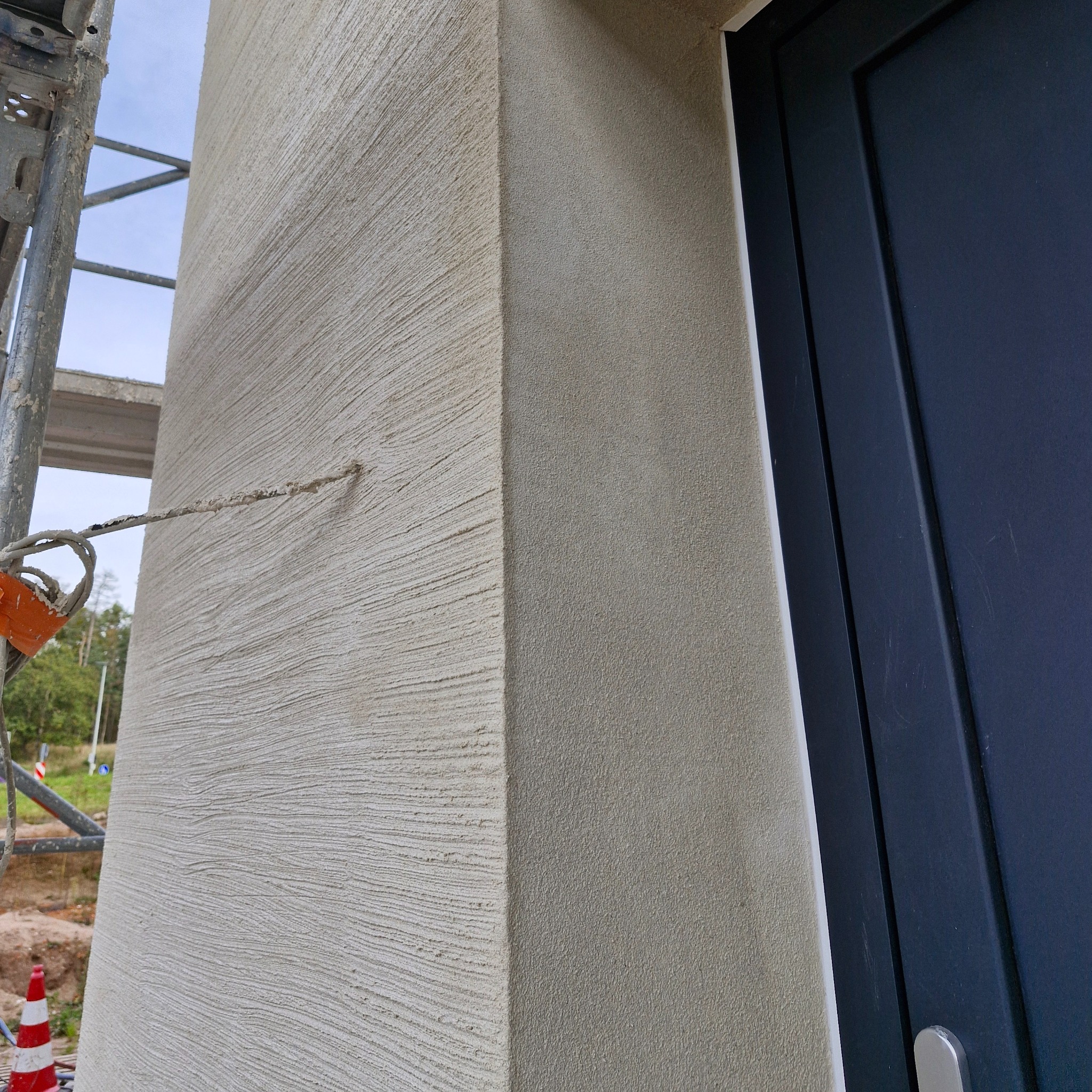 Fragment elewacji budynku z nałożonym tynkiem strukturalnym w odcieniach beżu, widoczny narożnik i fragment ościeżnicy drzwi, rusztowanie w tle.