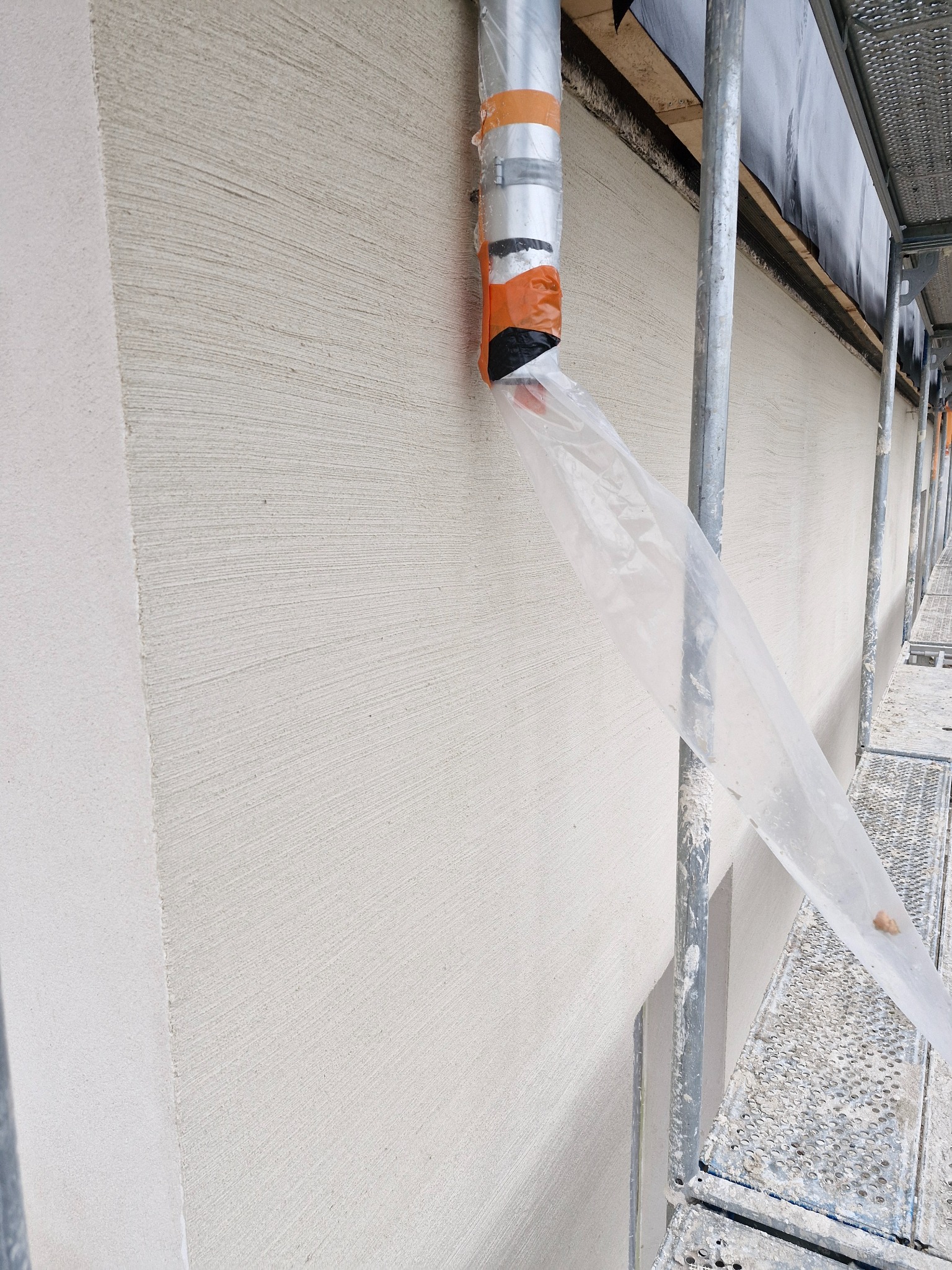 Świeżo nałożona, jasna struktura na elewacji budynku widoczna ze stojącego rusztowania, z rurą odprowadzającą pył przymocowaną do ściany taśmą pomarańczową.