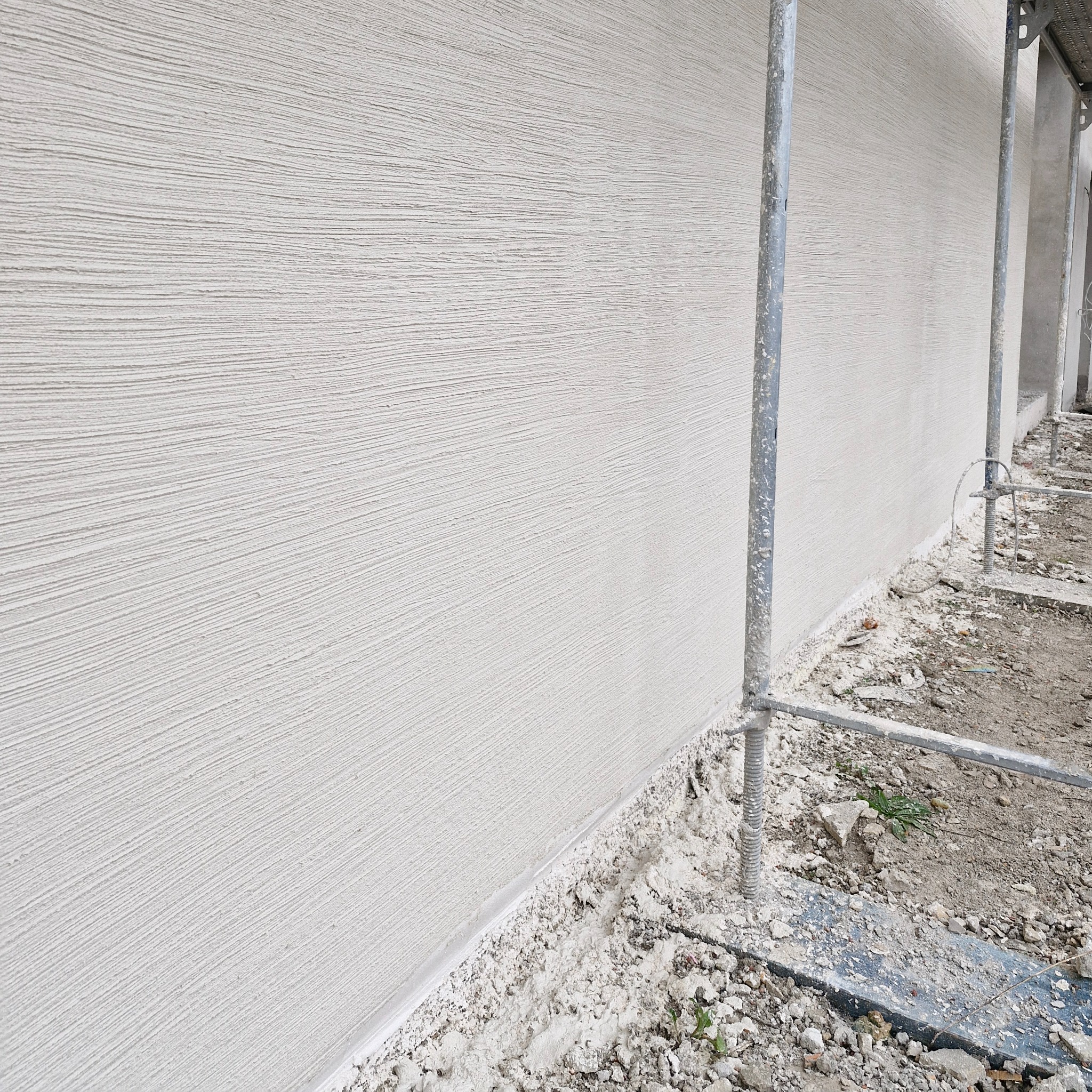 Świeżo nałożony, jasny tynk z wyraźną, poziomą fakturą na ścianie budynku, widoczne fragmenty metalowego rusztowania.