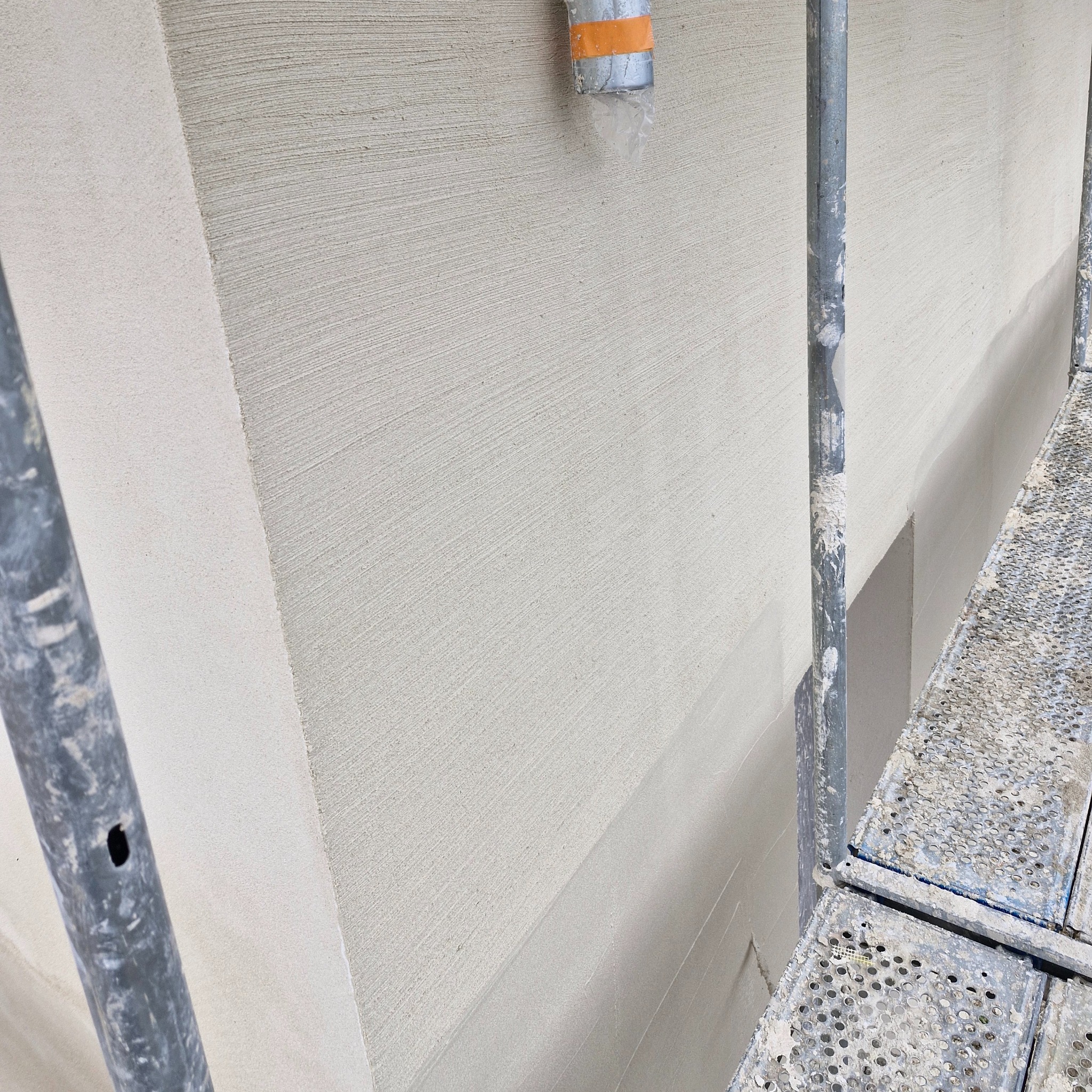 Świeżo nałożony, jasny tynk zewnętrzny z widoczną fakturą, fragment rusztowania z przodu, rura wentylacyjna przy górnej krawędzi ściany.