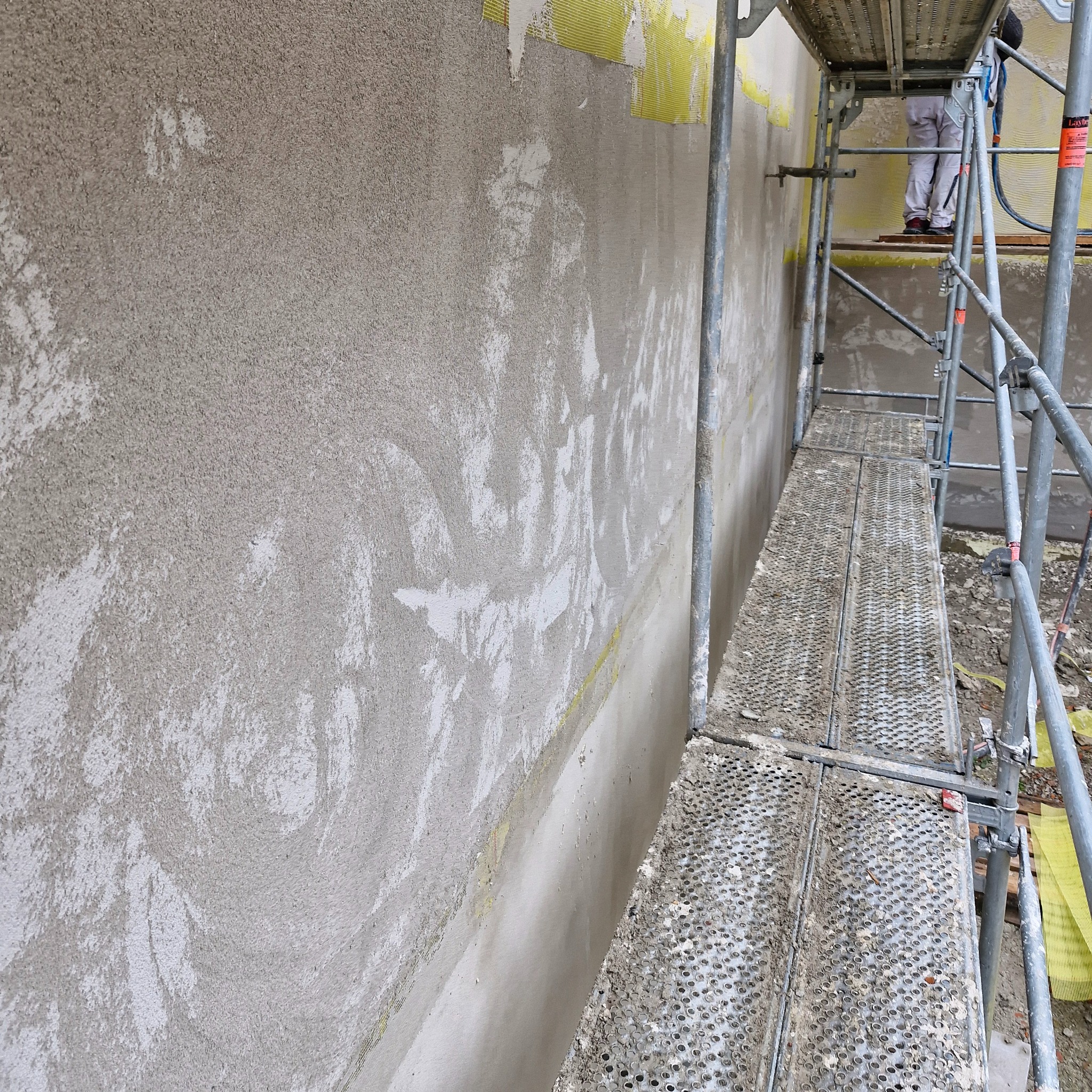Ściana budynku w trakcie nakładania tynku strukturalnego, widoczne fragmenty siatki wzmacniającej i rusztowanie, na którym stoi pracownik w kombinezonie.