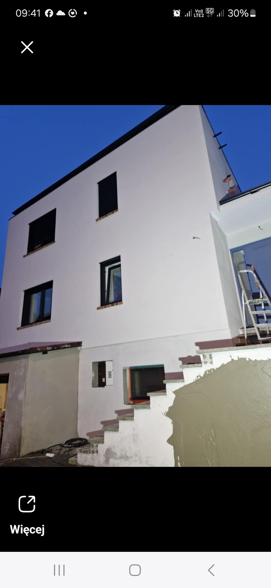 Dwupiętrowy budynek z białymi ścianami i czarnymi okiennicami w trakcie prac wykończeniowych; widoczne otynkowane schody wejściowe oraz drabina oparta o ścianę.