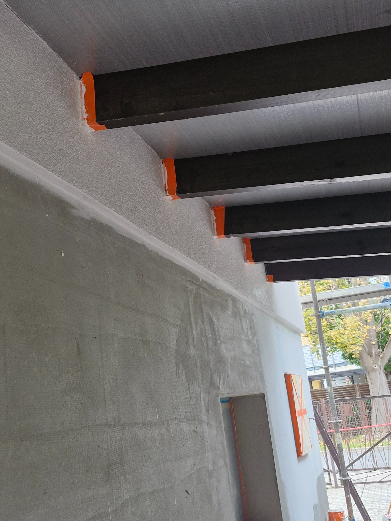 Wnętrze w trakcie prac wykończeniowych: surowa ściana z widoczną siatką, pomalowany pas pod sufitem, czarne belki stropowe z pomarańczowymi elementami mocującymi, widoczne rusztowanie i zarys okna.