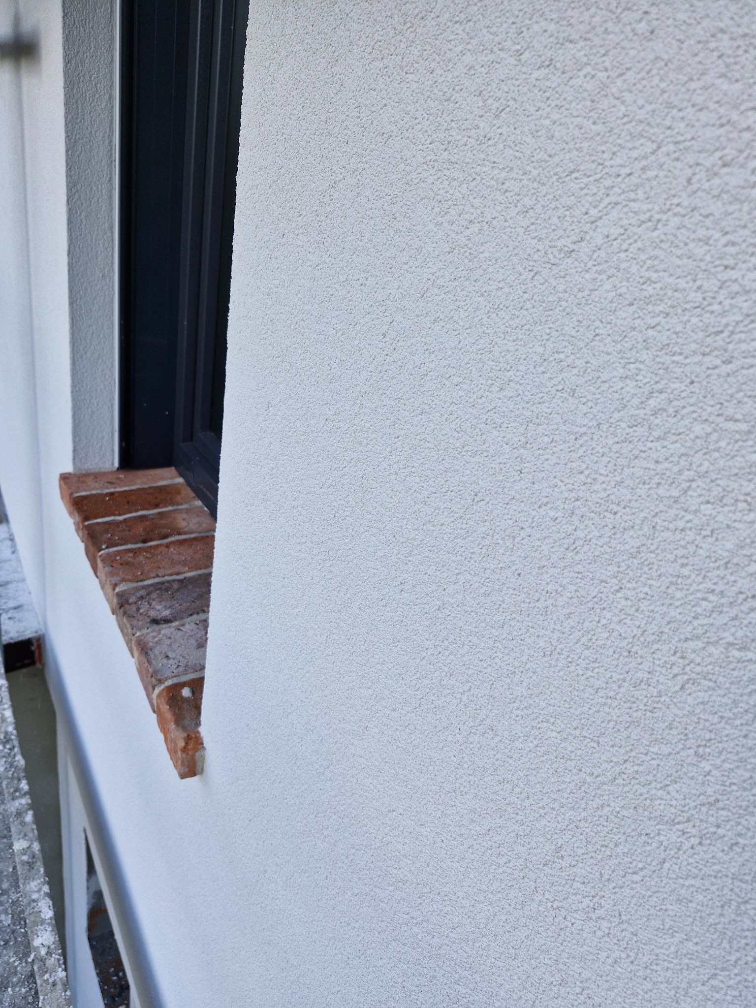 Fragment elewacji budynku po nałożeniu białego, baranka, widoczne obramowanie okna z ciemnego tworzywa i ceglany parapet.