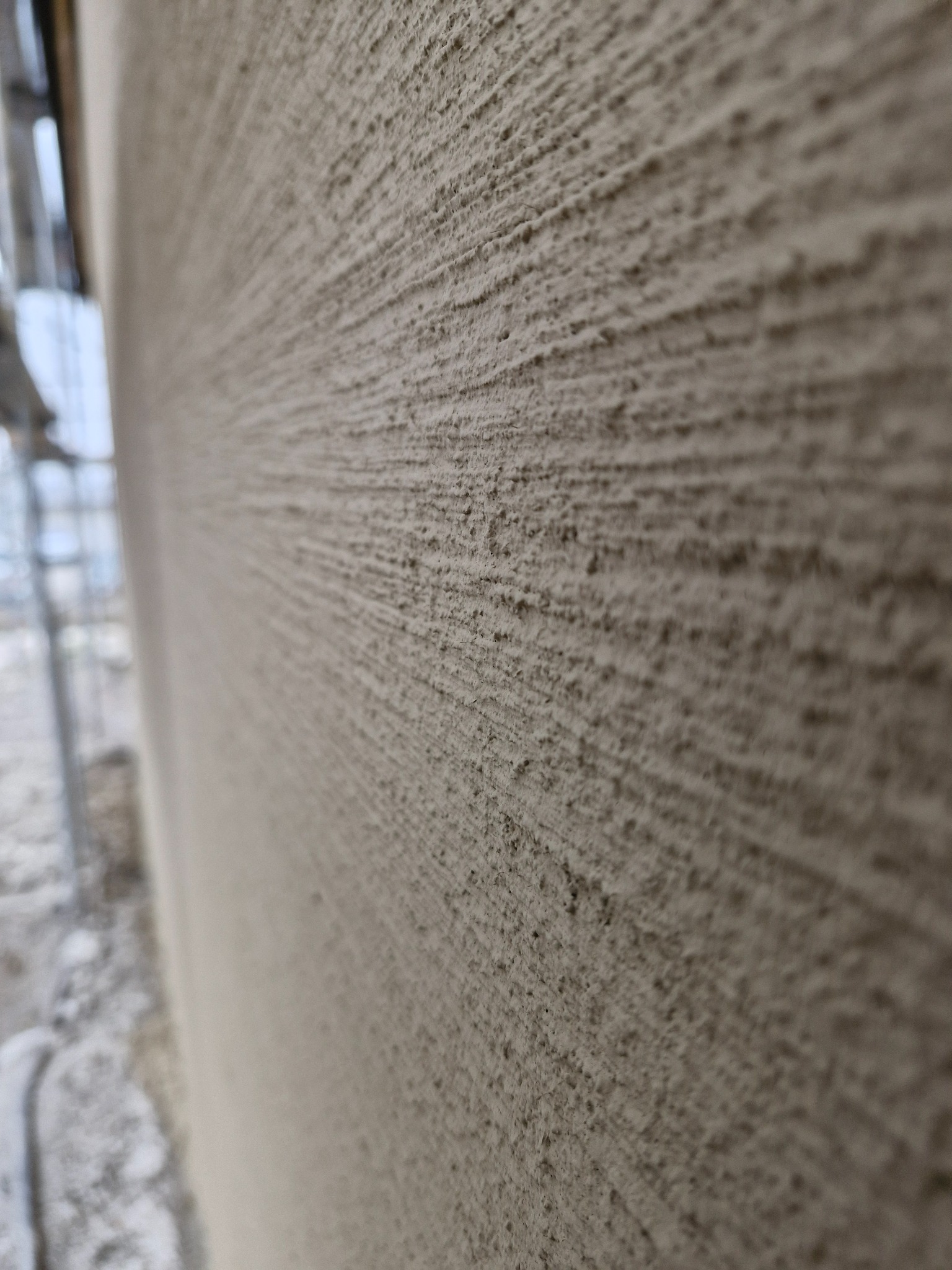 Szczegółowe ujęcie świeżo nałożonego, szarego tynku z wyraźnymi, poziomymi śladami po narzędziu, z fragmentem rusztowania widocznym w tle.