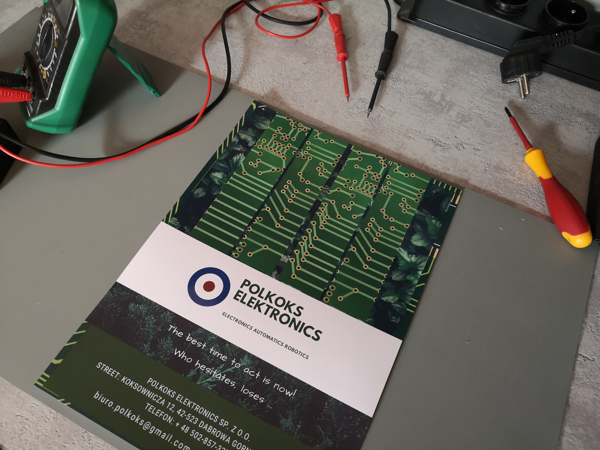 Wizytówka firmy Polkoks Elektronics z elementami obwodu drukowanego, leżąca obok multimetru, przewodów pomiarowych i śrubokręta na szarym tle.