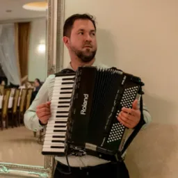 Mężczyzna z brodą w koszuli w kropki, trzymający czarny akordeon marki Roland FR-3X, stojący w sali balowej obok lustra w ozdobnej ramie.