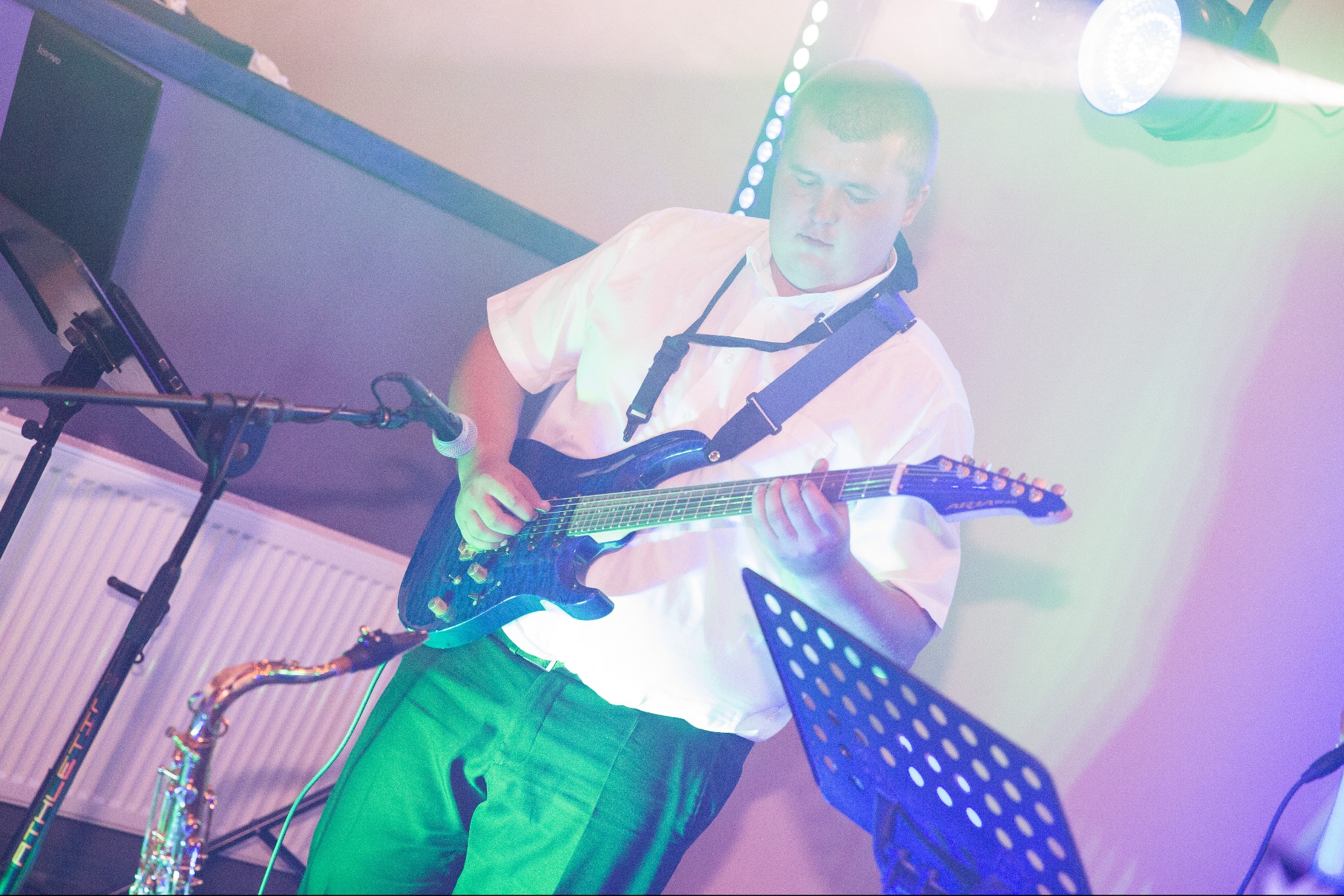 Gitarzysta w białej koszuli gra na niebieskiej gitarze elektrycznej podczas występu, obok widoczny saksofon i oświetlenie sceniczne.