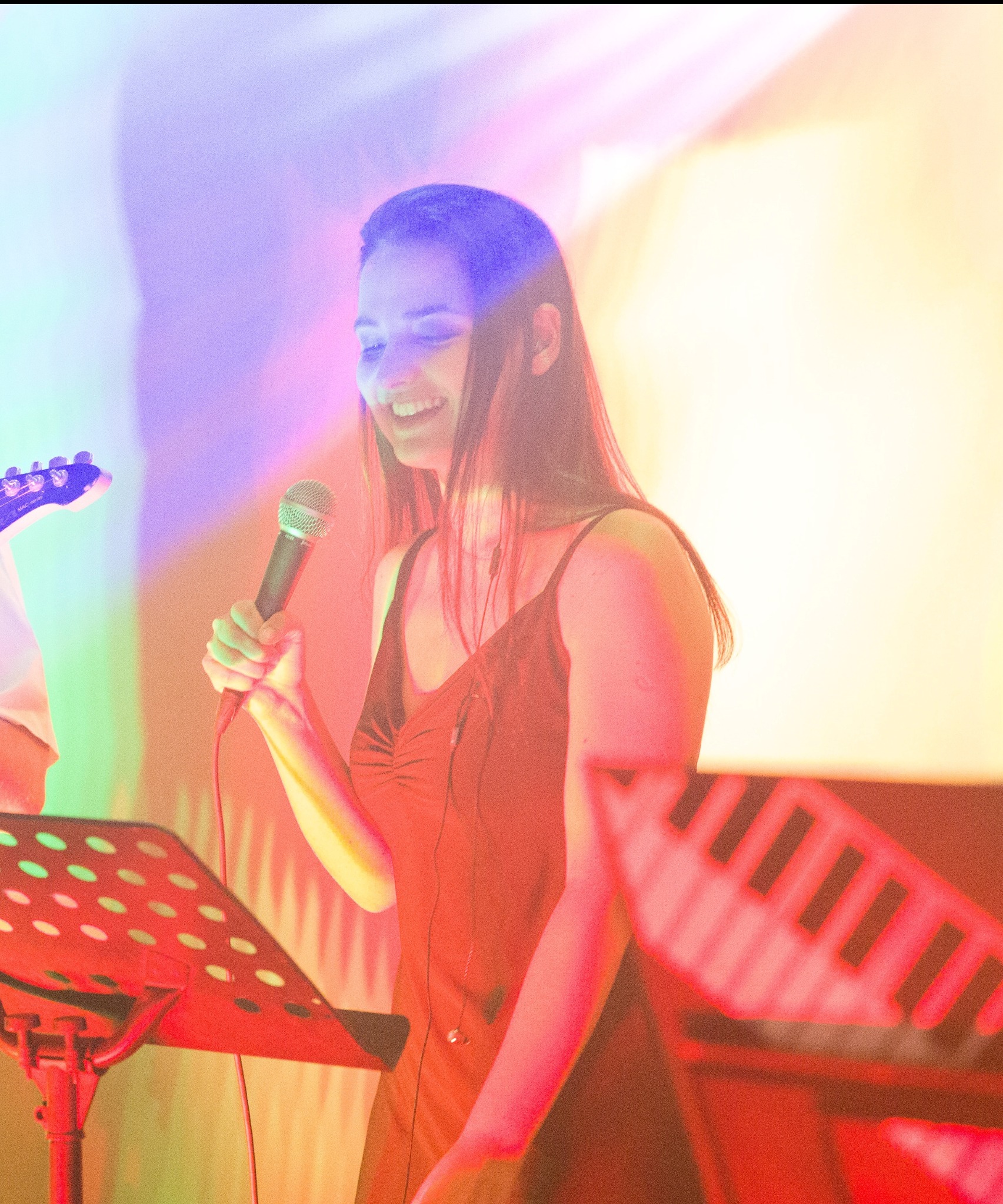 Uśmiechnięta wokalistka z mikrofonem podczas występu na scenie, z nutami na pulpicie i fragmentami klawiszy oraz gitary w tle, oświetlona kolorowymi światłami.