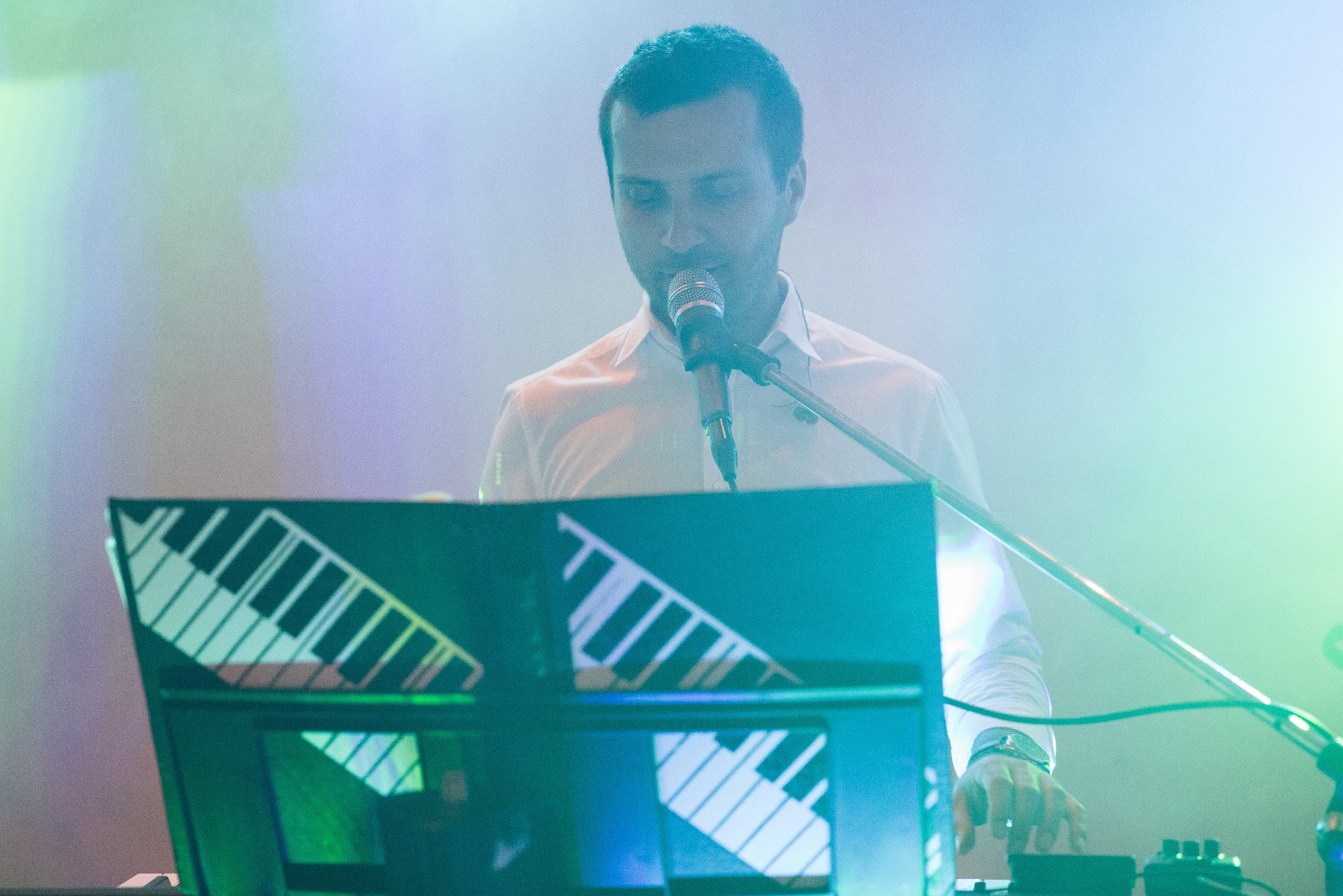 Mężczyzna w białej koszuli śpiewa do mikrofonu, grając na syntezatorze z graficznym przedstawieniem klawiatury na statywie w tle oświetlonym zielono-niebieskim światłem.