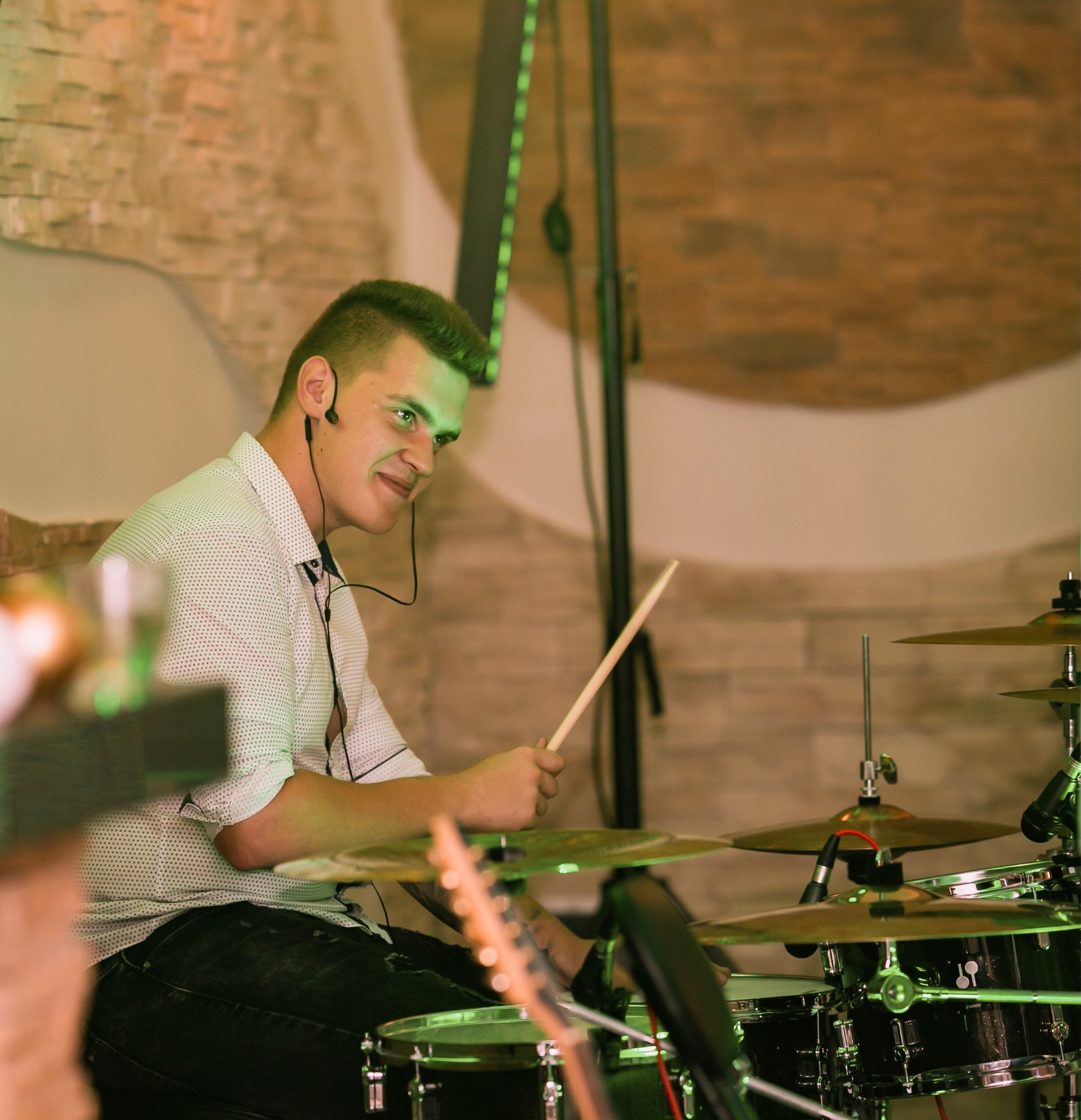 Młody perkusista z słuchawkami grający na perkusji podczas występu na scenie, oświetlony zielonym światłem.
