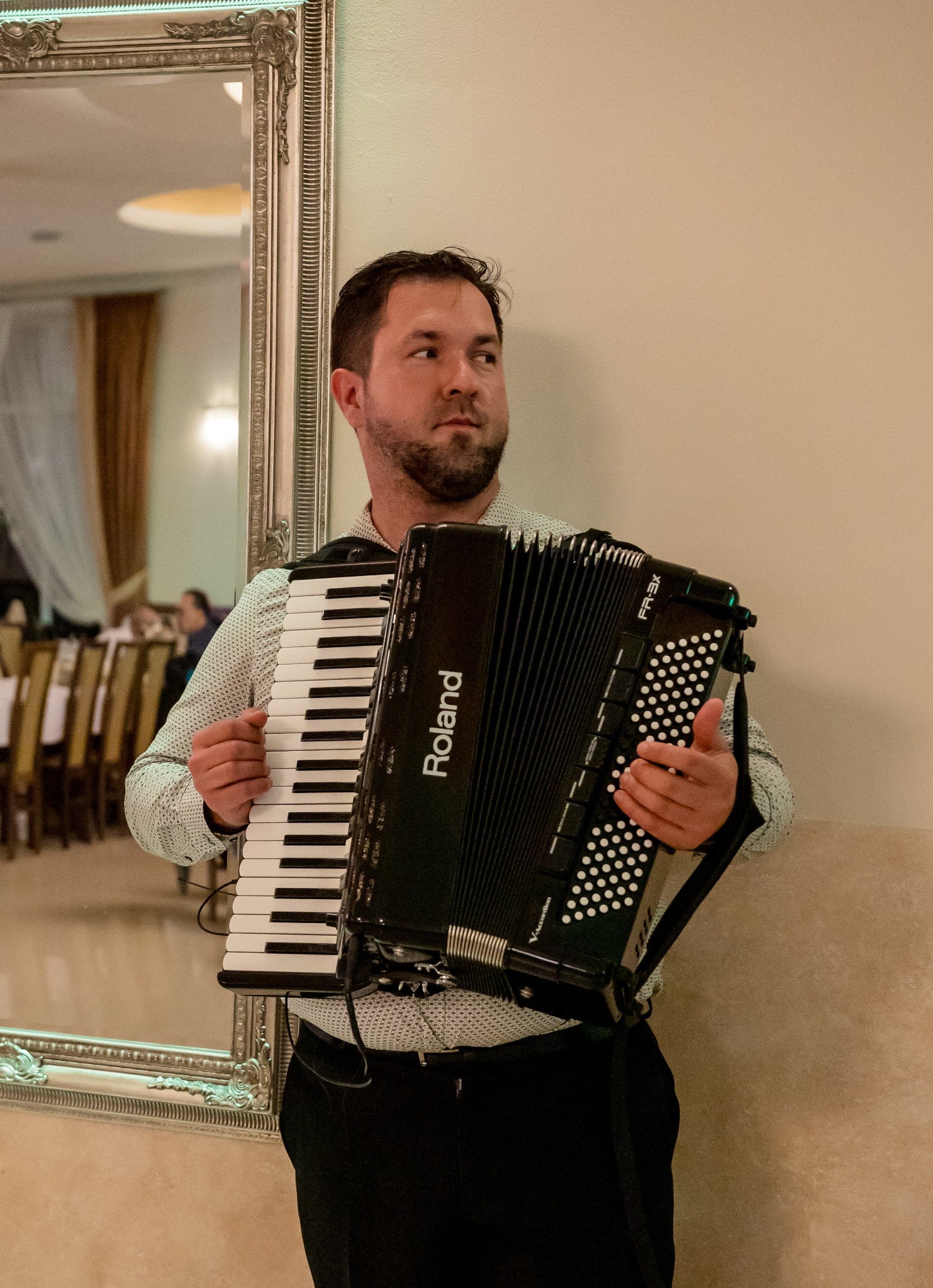 Mężczyzna z brodą w koszuli w kropki, trzymający czarny akordeon marki Roland FR-3X, stojący w sali balowej obok lustra w ozdobnej ramie.
