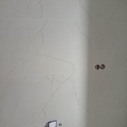 Nowobud Serhii Herasymenko - Ściana wyłożona białymi płytkami imitującymi marmur, z widocznymi otworami na gniazdka i zaklejonym otworem na rurę, ukazująca etap prac wykończeniowych.