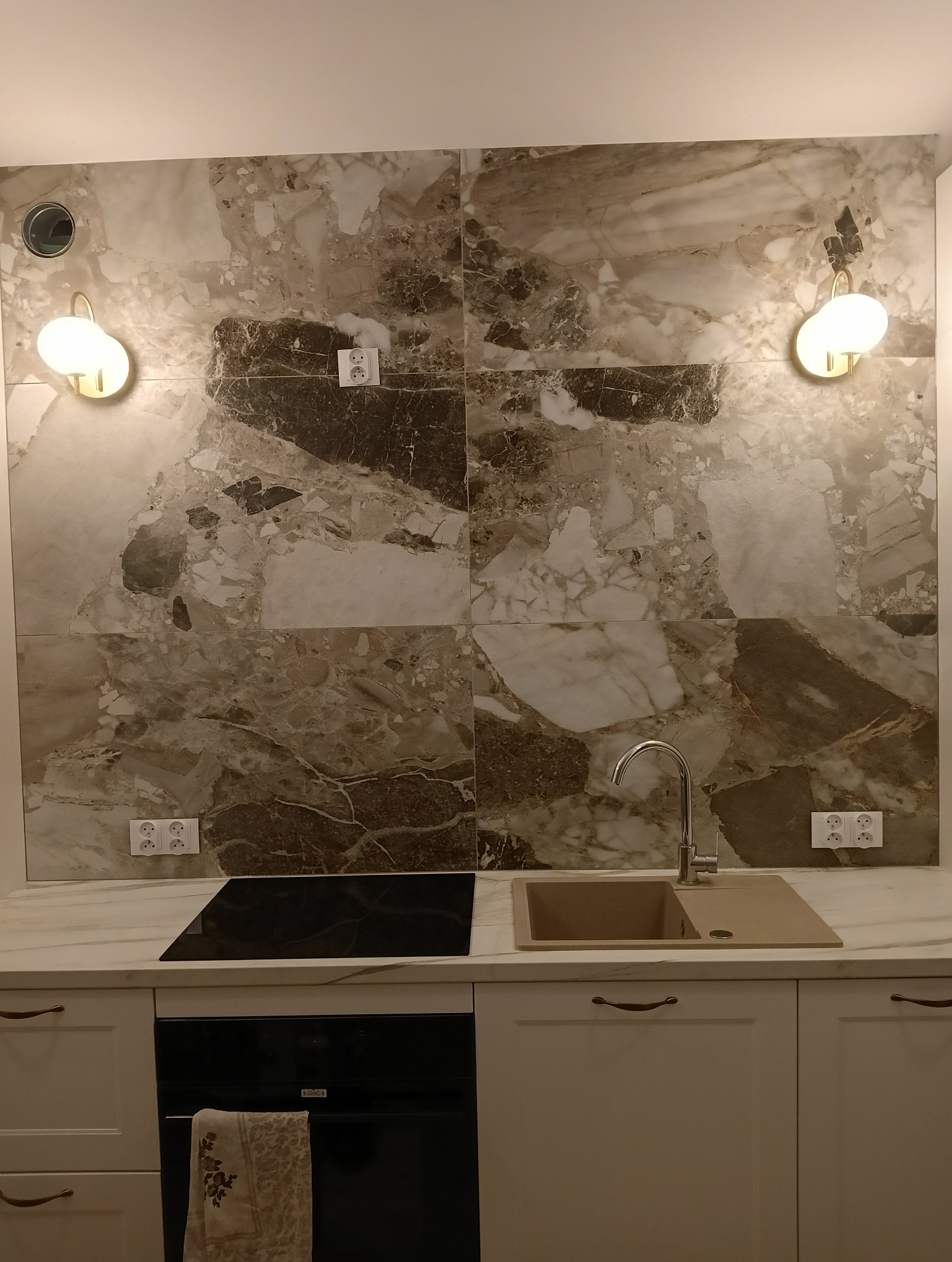 Nowoczesna kuchnia z marmurową ścianą, oświetleniem punktowym i beżowym zlewem. Widoczna płyta indukcyjna i piekarnik z ręcznikiem. Estetyczne wykończenie wnętrza.