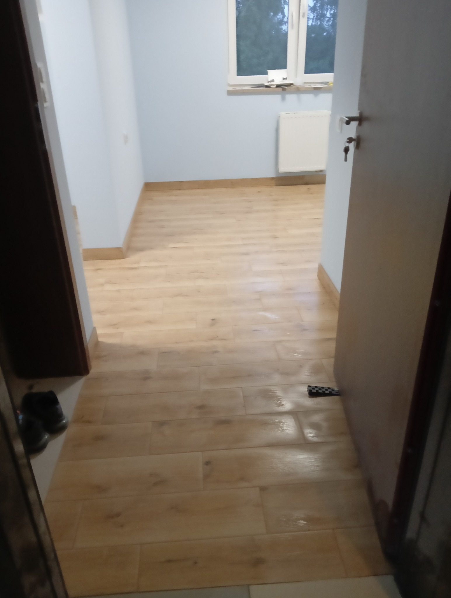 Ułożone panele podłogowe w jasnym odcieniu drewna w Kołobrzegu. Widok na wykończone wnętrze z oknem i grzejnikiem. Widoczny fragment otwartych drzwi.