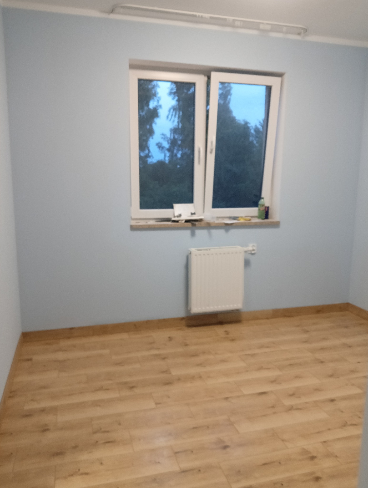 Jasny pokój po remoncie: ściany pomalowane na niebiesko, ułożone panele podłogowe w odcieniach brązu, biały grzejnik pod oknem. Na parapecie widoczne narzędzia.