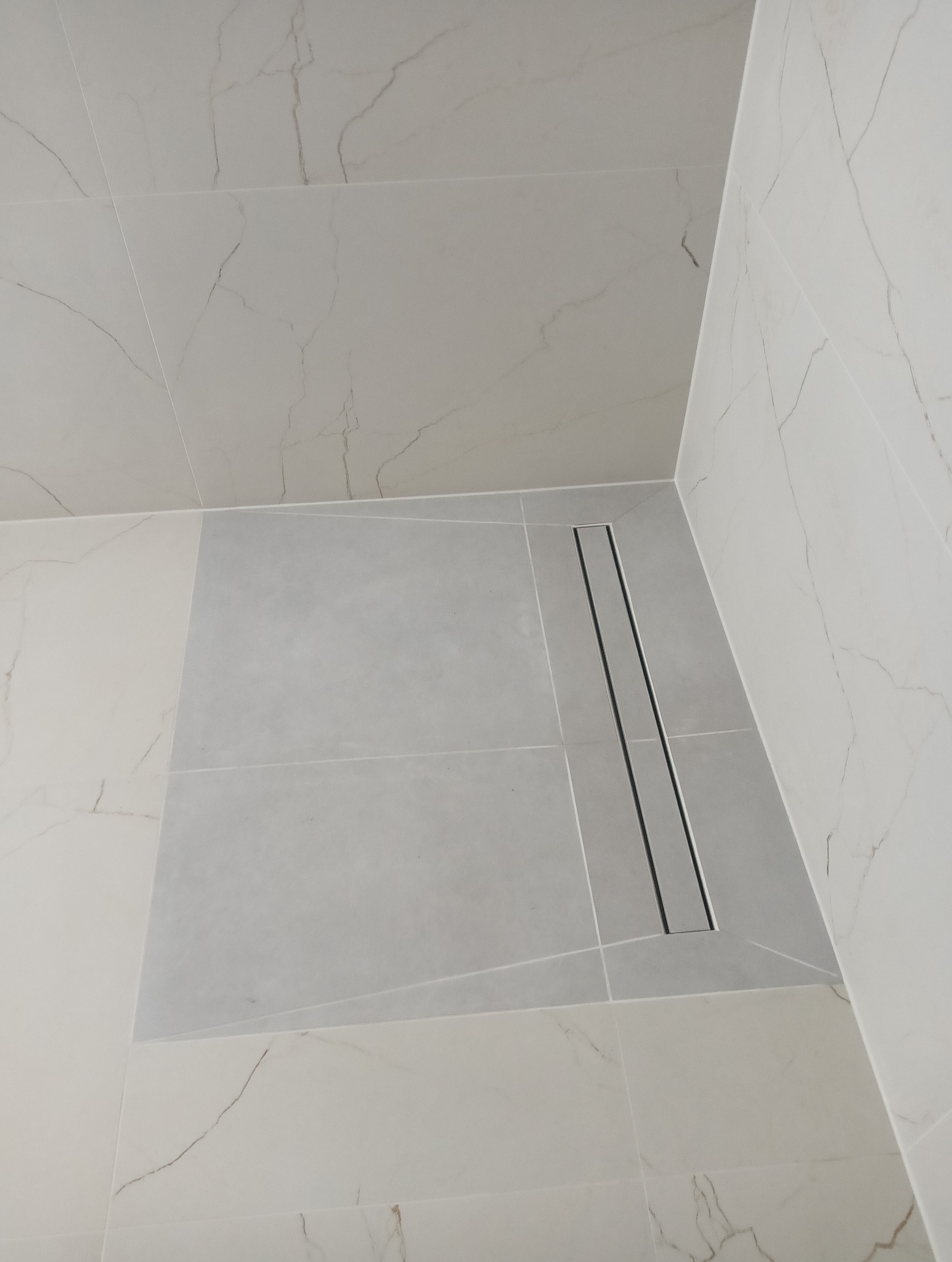 Wykończona strefa prysznicowa z odpływem liniowym i płytkami imitującymi marmur. Minimalistyczny design łazienki z szarymi i białymi płytkami na ścianach i podłodze.
