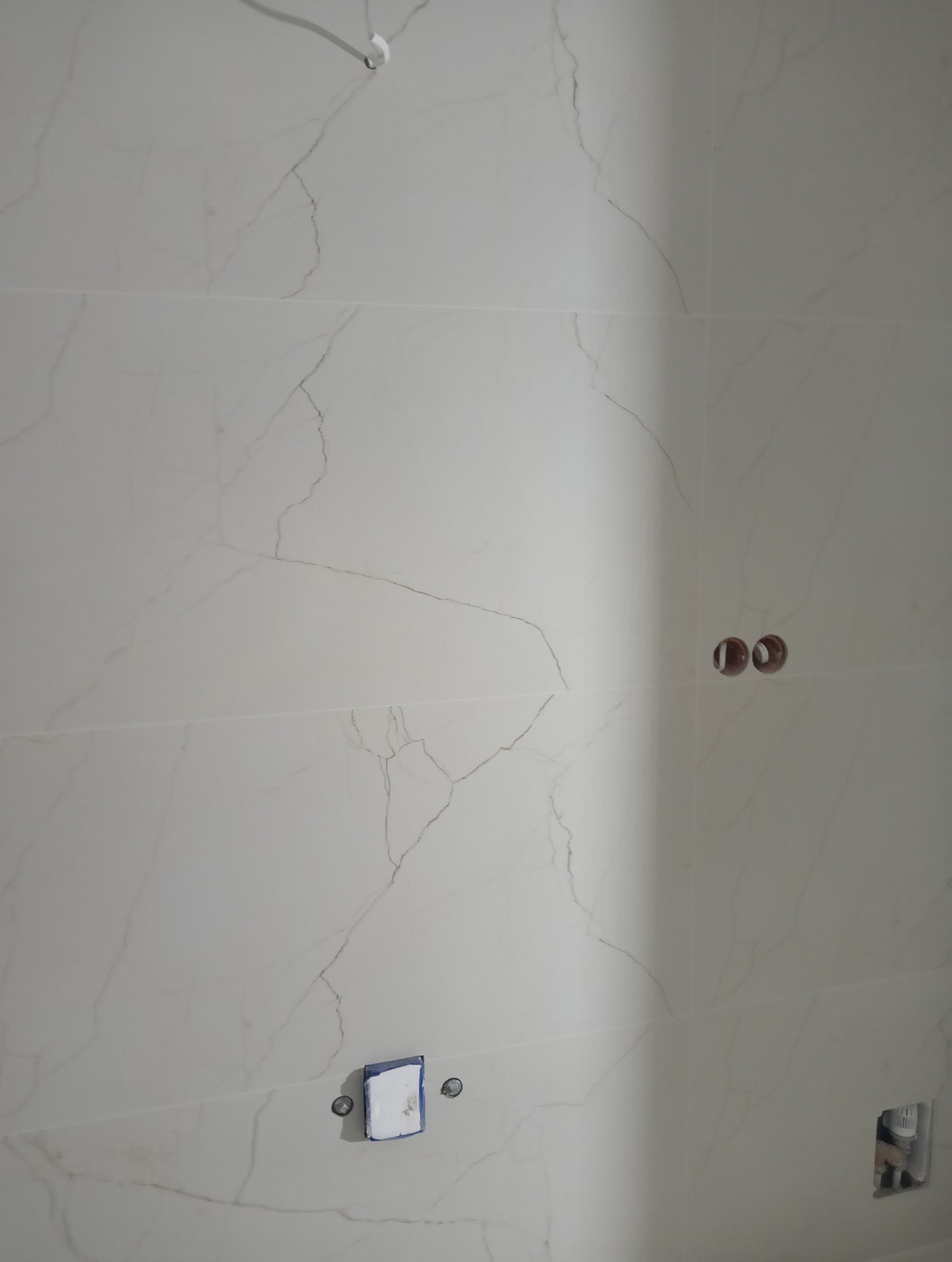 Ściana wyłożona białymi płytkami imitującymi marmur, z widocznymi otworami na gniazdka i zaklejonym otworem na rurę, ukazująca etap prac wykończeniowych.