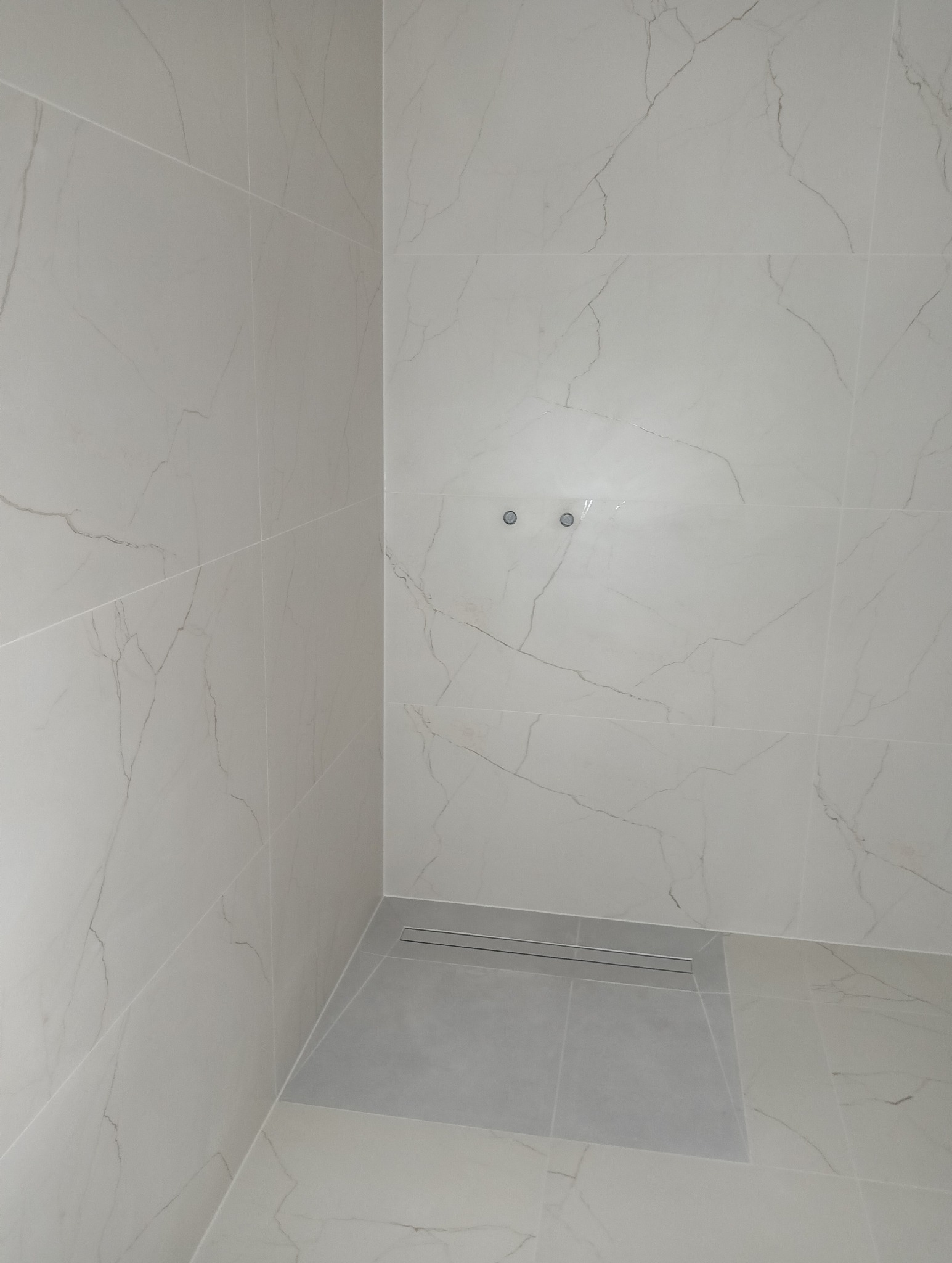 Wykończona kabina prysznicowa z białymi płytkami imitującymi marmur i odpływem liniowym. Widoczne dwa otwory na armaturę. Minimalistyczny design.