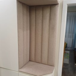 Krzysztof Szliselman - Beżowa, tapicerowana wnęka siedziskowa w zabudowie meblowej. Pionowe przeszycia na oparciu, miękkie siedzisko. Białe fronty szafek, widok na korytarz.