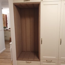 Krzysztof Szliselman - Biała zabudowa meblowa z tapicerowanym, brązowym siedziskiem wnękowym, widok z boku. Złote uchwyty dodają elegancji. W tle widoczna kuchnia.