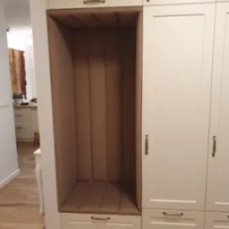 Biała zabudowa meblowa z tapicerowanym, brązowym siedziskiem wnękowym, widok z boku. Złote uchwyty dodają elegancji. W tle widoczna kuchnia.