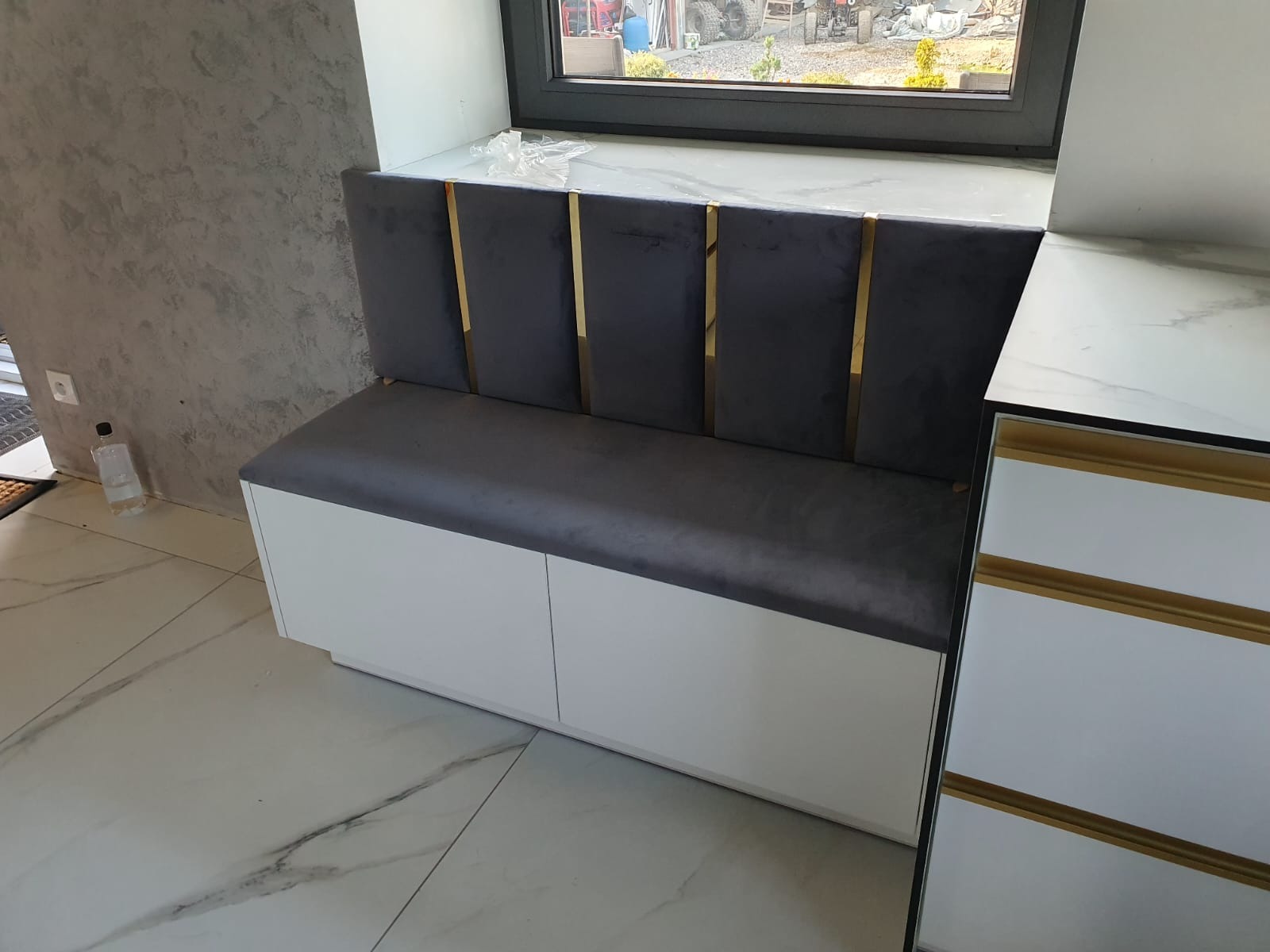 Szara, tapicerowana ławka ze złotymi akcentami, wkomponowana pod oknem. Białe szafki pod siedziskiem, marmurowe blaty i podłoga. Nowoczesny design.