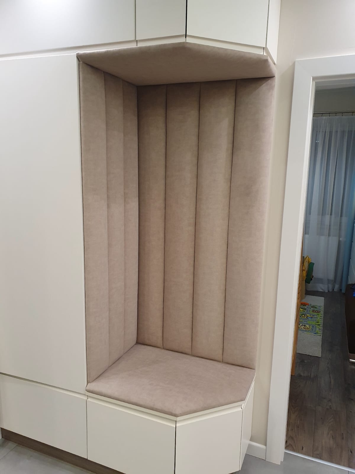 Beżowa, tapicerowana wnęka siedziskowa w zabudowie meblowej. Pionowe przeszycia na oparciu, miękkie siedzisko. Białe fronty szafek, widok na korytarz.