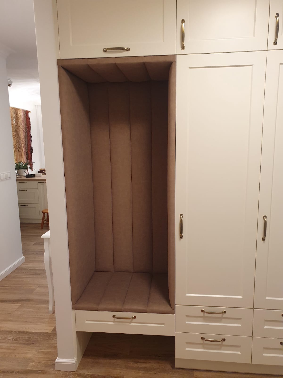 Biała zabudowa meblowa z tapicerowanym, brązowym siedziskiem wnękowym, widok z boku. Złote uchwyty dodają elegancji. W tle widoczna kuchnia.