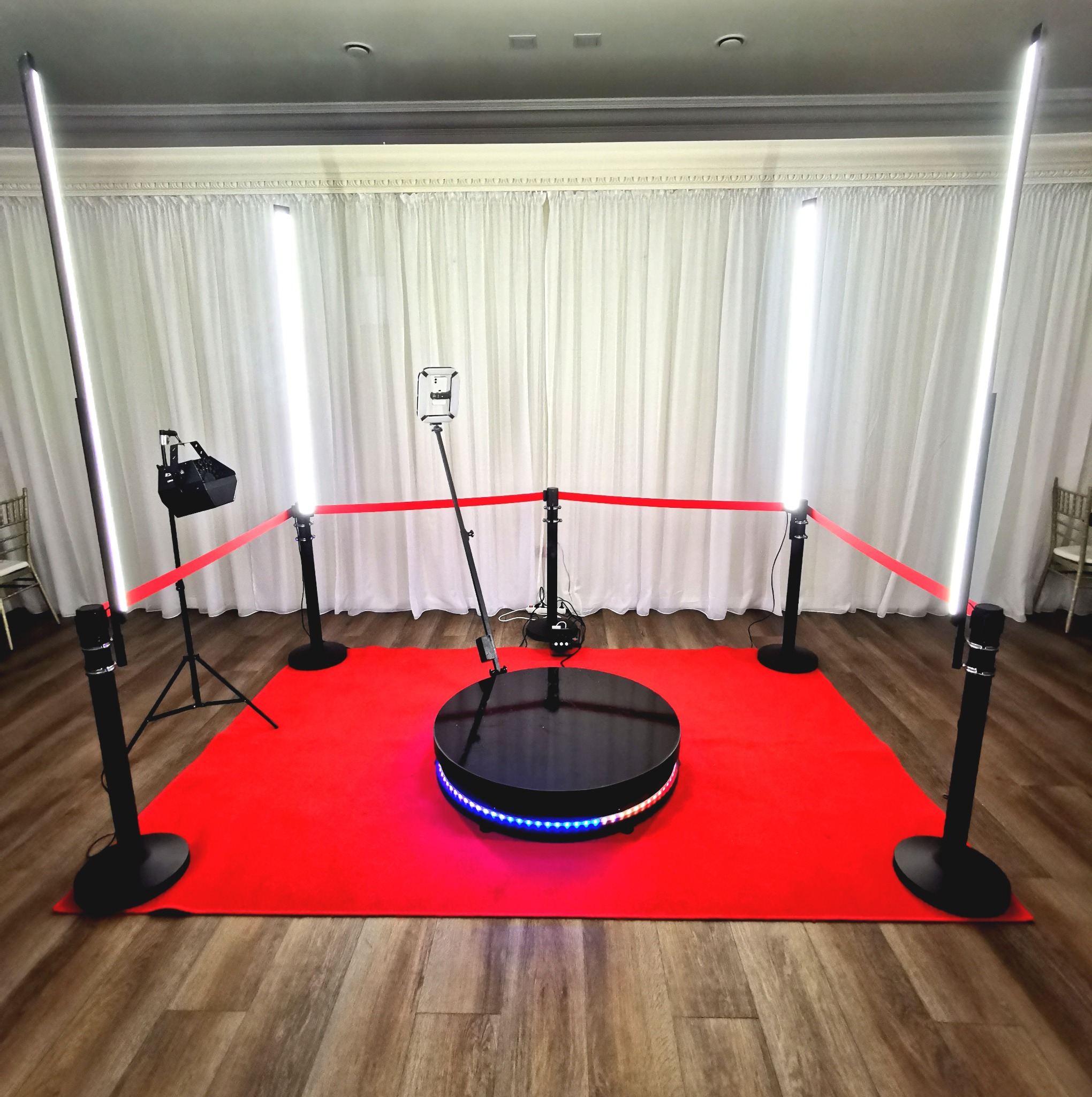 Okrągła platforma z podświetleniem LED otoczona czerwonym dywanem i taśmą, oświetlona lampami LED, ustawiona na drewnianej podłodze przed białymi zasłonami.