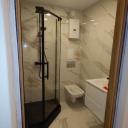 Aqfast - Nowoczesna łazienka z kabiną prysznicową w czarnej ramie, marmurowe płytki na ścianach i podłodze, biała umywalka z szafką i bidet, widok zza framugi drzwi.