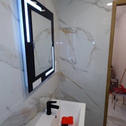 Aqfast - Biała łazienka z marmurowymi płytkami, podświetlanym lustrem w czarnej ramie, czarną baterią umywalkową i narzędziami w umywalce, widoczne niezaślepione kable wystające ze ściany.