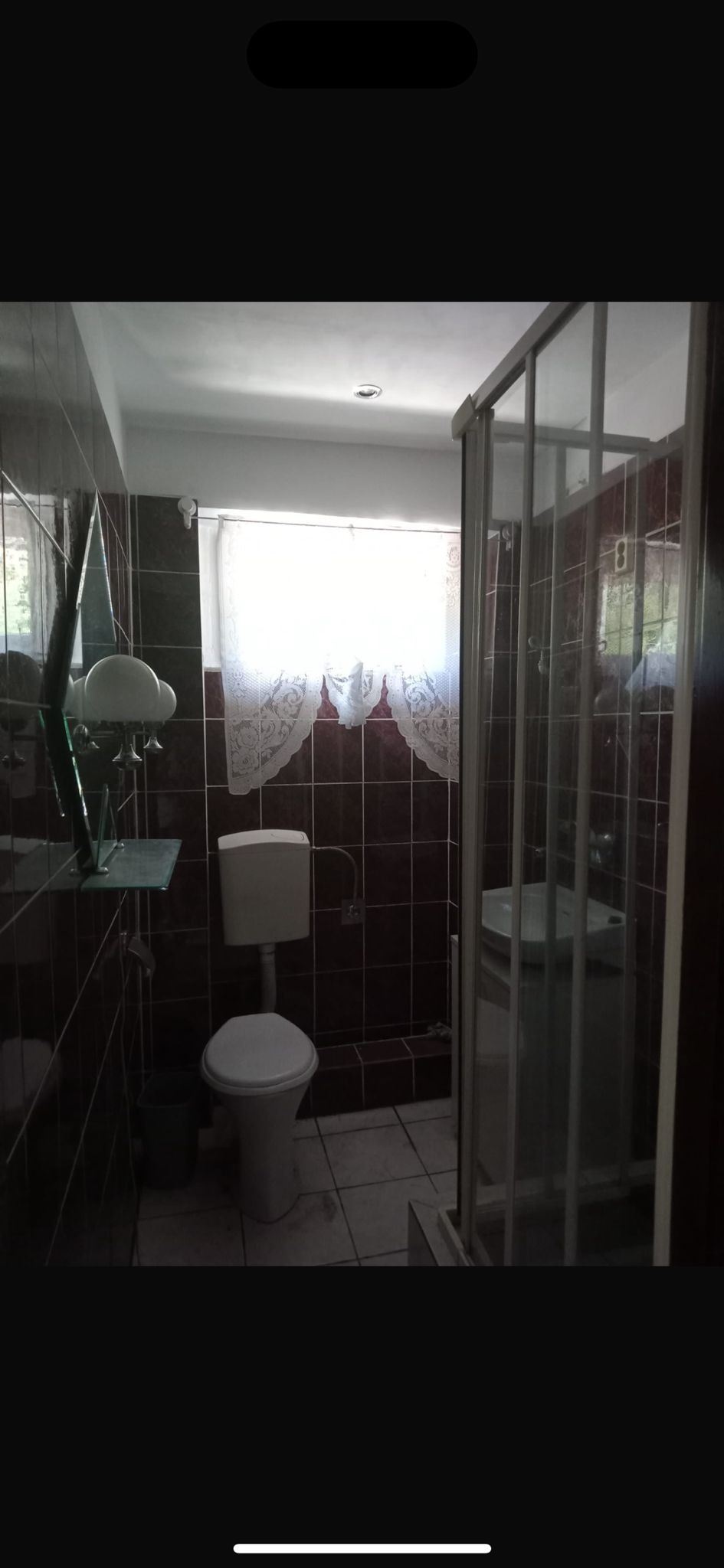 Ciemna łazienka z brązowymi płytkami, kabiną prysznicową ze srebrną ramą, toaletą i ozdobną lampą na szklanej półce.