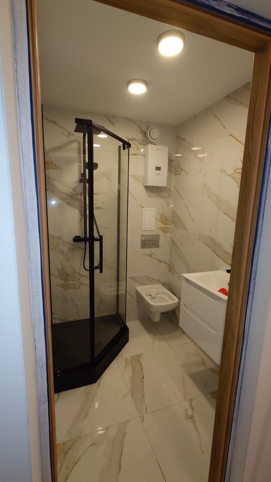 Nowoczesna łazienka z kabiną prysznicową w czarnej ramie, marmurowe płytki na ścianach i podłodze, biała umywalka z szafką i bidet, widok zza framugi drzwi.