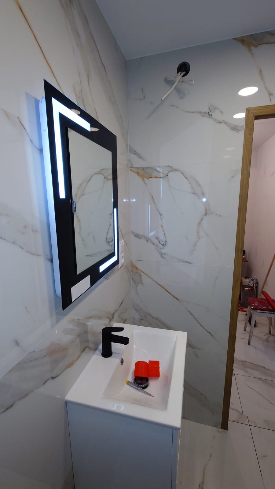 Biała łazienka z marmurowymi płytkami, podświetlanym lustrem w czarnej ramie, czarną baterią umywalkową i narzędziami w umywalce, widoczne niezaślepione kable wystające ze ściany.