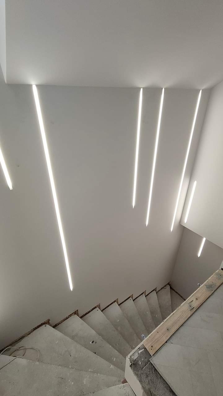 Wnętrze klatki schodowej w trakcie prac wykończeniowych, widoczne surowe betonowe schody i nowoczesne oświetlenie LED wpuszczone w gładką, białą ścianę i sufit.