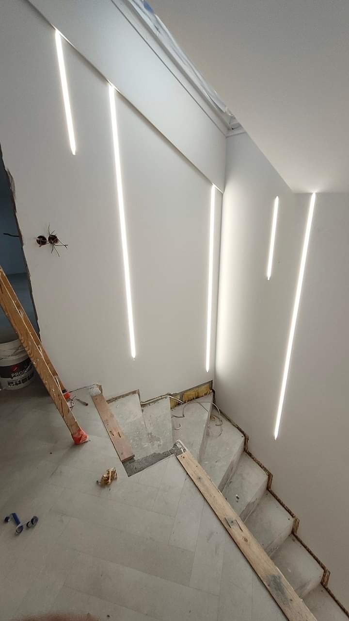 Klatka schodowa w trakcie remontu, widoczne surowe schody zabezpieczone deskami, ściany z pionowymi wpuszczonymi pasami oświetlenia LED, puszki elektryczne bez osprzętu.