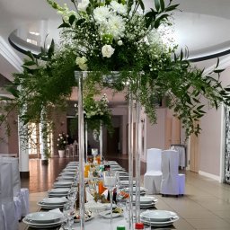 MAGIC PARTY Karolina Stolarek - Uroczysta sala bankietowa z długim stołem udekorowanym białymi kwiatami i zielonymi liśćmi w wysokich, przezroczystych wazonach, zastawionym talerzami, szklankami i napojami.