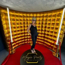 Kobieta w czarnym kombinezonie i koronie na głowie pozuje na obrotowej platformie z napisem Magic Party Karolina Stolarek, na tle złotej dmuchanej ściany i czerwonego dywanu z oświetleniem.
