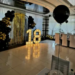 Dekoracja sali na 18 urodziny: podświetlane cyfry '18', złote frędzle, girlanda z czarnych i złotych balonów oraz duży czarny balon na sali z nakrytymi stołami.