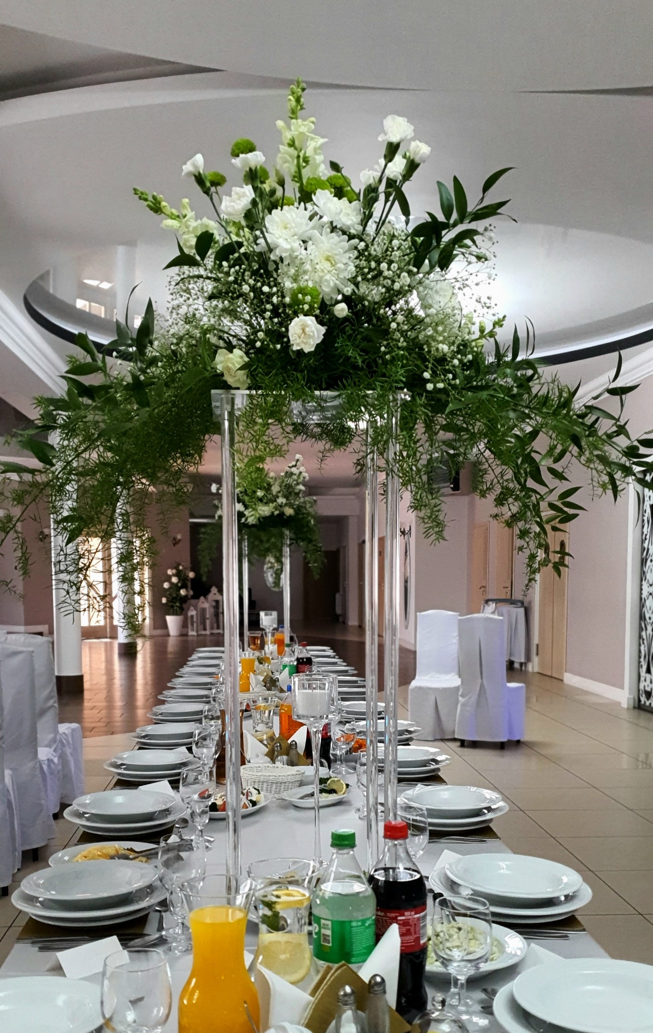 Uroczysta sala bankietowa z długim stołem udekorowanym białymi kwiatami i zielonymi liśćmi w wysokich, przezroczystych wazonach, zastawionym talerzami, szklankami i napojami.