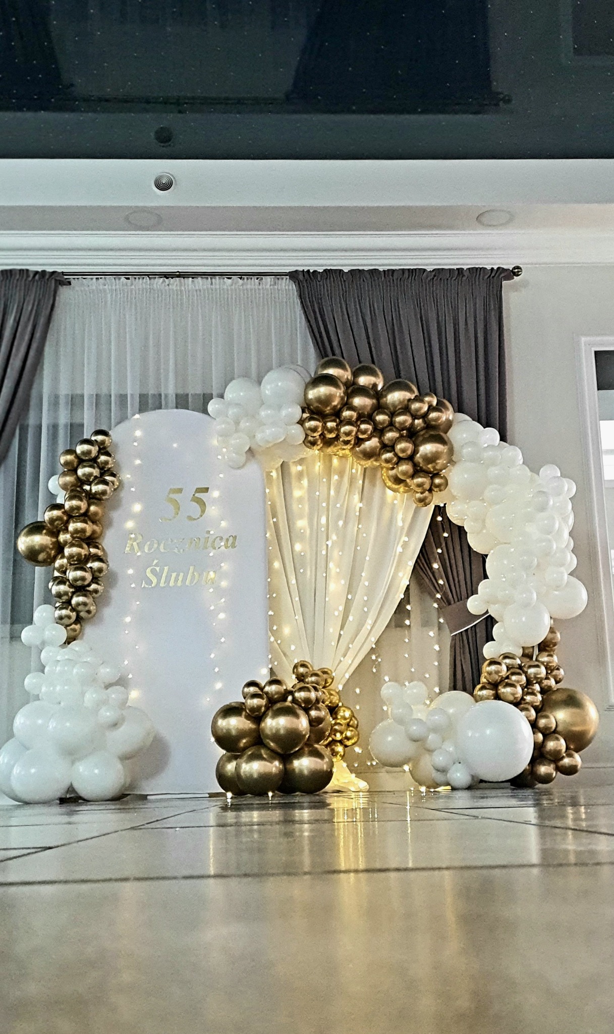 Dekoracja na 55. Rocznicę Ślubu z białych i złotych balonów, podświetlona girlandą lampek, na tle białej ścianki i drapowanych zasłon.