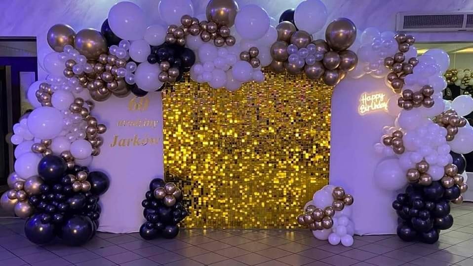 Dekoracja urodzinowa z błyszczącym złotym tłem, balonami w kolorach białym, czarnym i złotym oraz napisem neonowym 'Happy Birthday'.
