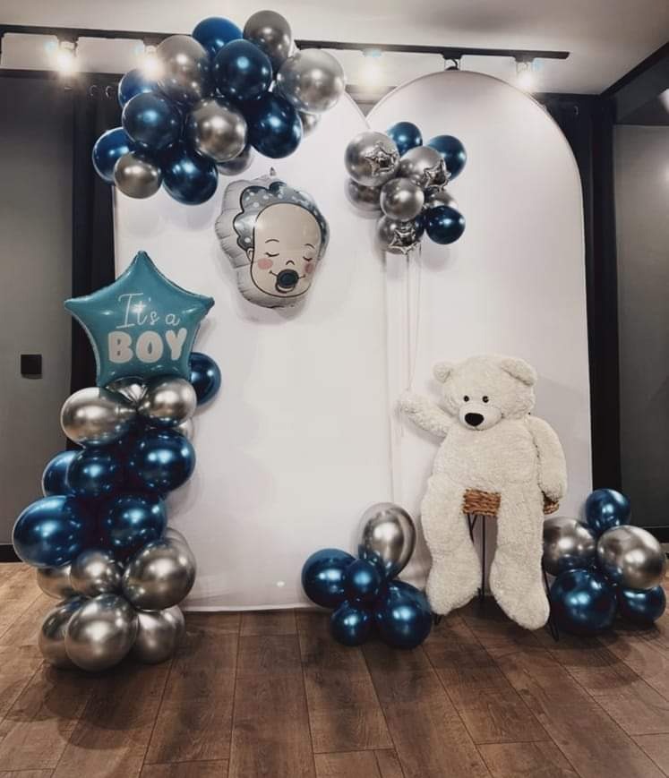 Dekoracja balonowa w odcieniach niebieskiego i srebrnego z pluszowym misiem na przyjęcie z okazji narodzin syna, ustawiona na drewnianej podłodze.