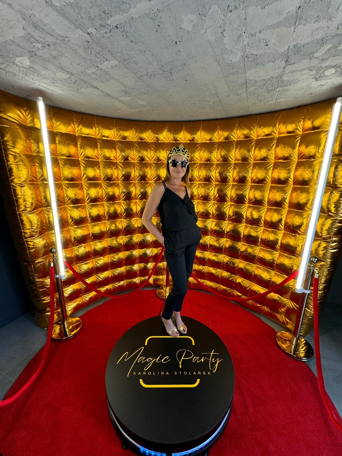 Kobieta w czarnym kombinezonie i koronie na głowie pozuje na obrotowej platformie z napisem Magic Party Karolina Stolarek, na tle złotej dmuchanej ściany i czerwonego dywanu z oświetleniem.